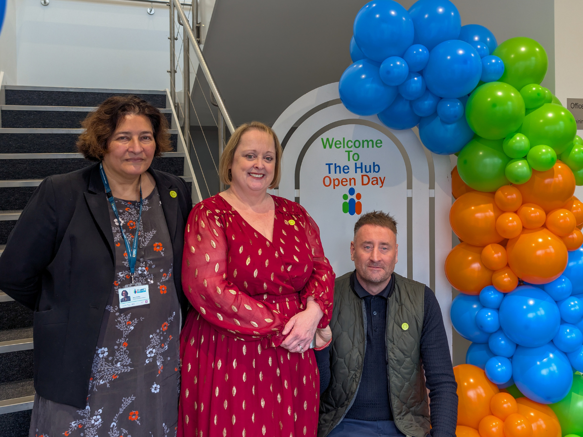 Three people in front of a sign (with balloon arch) that says Welcome to the hub open day